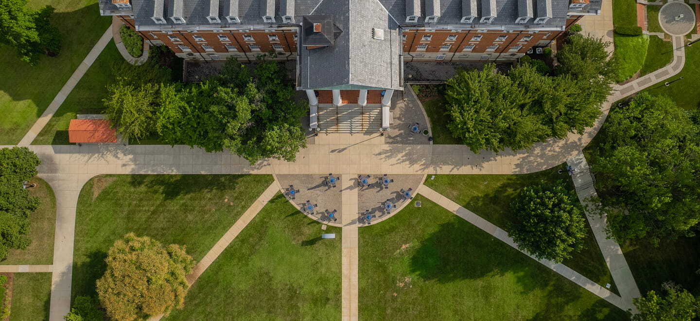 Aerial view of a brick academic building with people gathered at the entrance, surrounded by sidewalks, grass, and trees on a college campus.