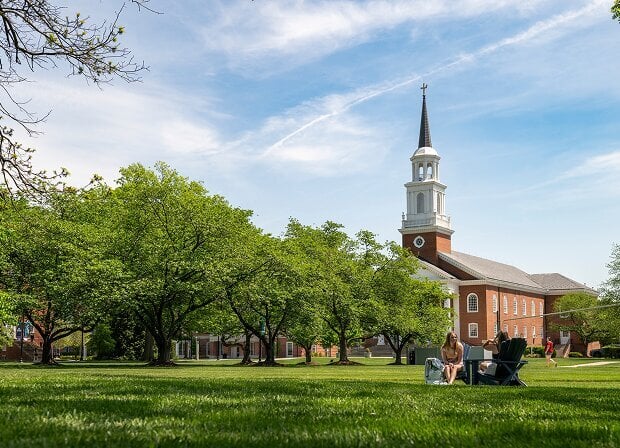 A grassy campus lawn with people sitting and a red-brick building with a white steeple in the background under a blue sky.